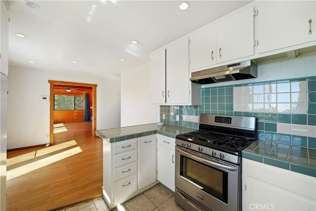 kitchen with tile countertops, stainless steel gas range, a peninsula, under cabinet range hood, and white cabinetry