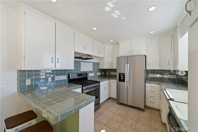 kitchen with tile countertops, stainless steel appliances, white cabinets, a peninsula, and under cabinet range hood