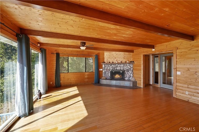 unfurnished living room featuring hardwood / wood-style floors, beamed ceiling, a stone fireplace, wooden walls, and a ceiling fan