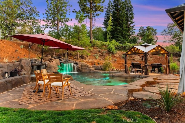 pool at dusk with a gazebo, grilling area, pool water feature, and a patio