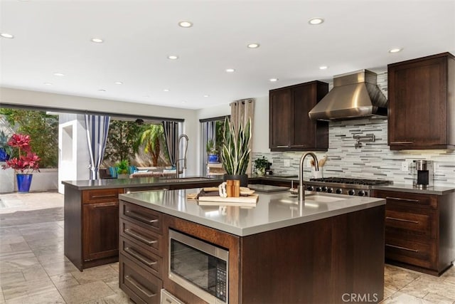 kitchen featuring a center island with sink, tasteful backsplash, appliances with stainless steel finishes, a sink, and wall chimney exhaust hood