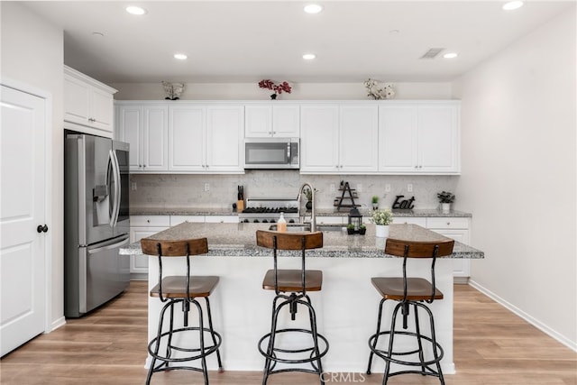 kitchen featuring light wood-type flooring, stainless steel appliances, white cabinets, light stone counters, and a kitchen island with sink