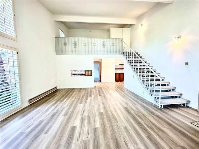 unfurnished living room featuring baseboard heating, beam ceiling, light hardwood / wood-style floors, and a high ceiling