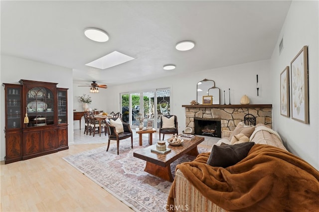 living room with a skylight, ceiling fan, a stone fireplace, and light hardwood / wood-style floors