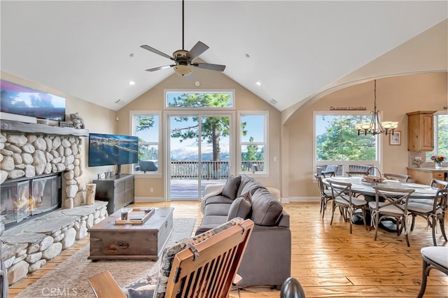 living room featuring ceiling fan with notable chandelier, high vaulted ceiling, light wood-type flooring, and a stone fireplace