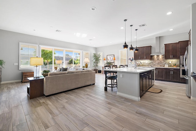 kitchen featuring a kitchen breakfast bar, wall chimney range hood, light hardwood / wood-style flooring, hanging light fixtures, and an island with sink