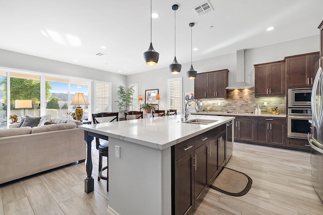 kitchen featuring a breakfast bar, wall chimney range hood, sink, an island with sink, and appliances with stainless steel finishes