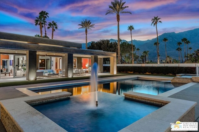 pool at dusk with an in ground hot tub, a patio, a mountain view, and an outdoor hangout area