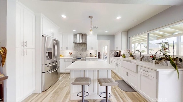 kitchen with stainless steel appliances, wall chimney range hood, light hardwood / wood-style floors, decorative light fixtures, and white cabinets