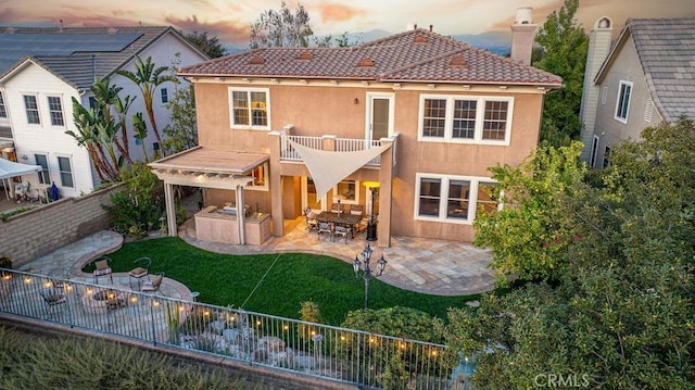 back house at dusk with an outdoor kitchen, a balcony, a patio, and a lawn