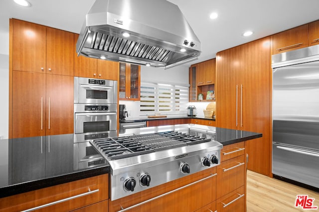 kitchen featuring dark stone counters, light hardwood / wood-style flooring, exhaust hood, and appliances with stainless steel finishes