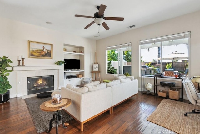 living room featuring dark hardwood / wood-style flooring, built in features, ceiling fan, and a tiled fireplace