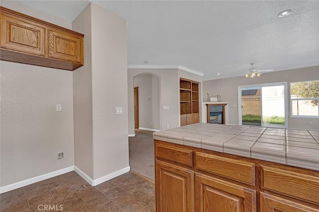 kitchen featuring arched walkways, a glass covered fireplace, tile counters, and baseboards