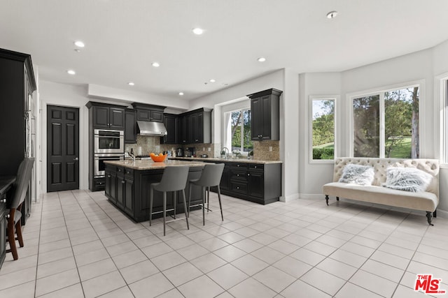 kitchen with light stone countertops, tasteful backsplash, a breakfast bar, stainless steel double oven, and an island with sink