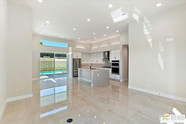 kitchen with wall chimney exhaust hood, a center island with sink, sink, appliances with stainless steel finishes, and white cabinetry