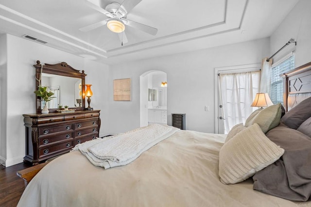 bedroom with dark wood-type flooring, connected bathroom, ceiling fan, and a tray ceiling