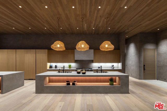 kitchen featuring an island with sink, wall chimney exhaust hood, sink, light hardwood / wood-style floors, and hanging light fixtures