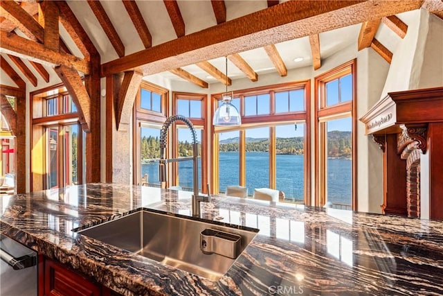 kitchen with dark stone countertops, beam ceiling, sink, hanging light fixtures, and a water view