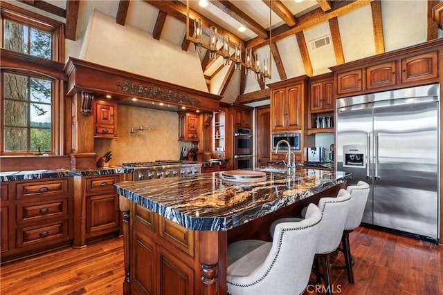 kitchen featuring a kitchen island with sink, high vaulted ceiling, dark stone counters, and built in appliances