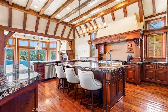 kitchen with pendant lighting, dishwasher, beamed ceiling, dark stone countertops, and a kitchen island with sink