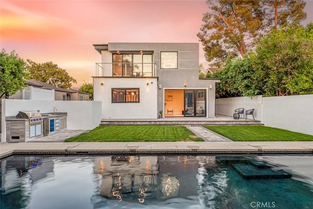 back house at dusk with a fenced in pool, a balcony, a yard, exterior kitchen, and a patio