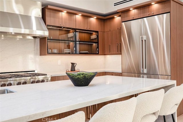 kitchen featuring light stone countertops, a kitchen bar, wall chimney range hood, and appliances with stainless steel finishes