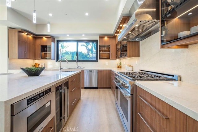 kitchen featuring wall chimney range hood, sink, light stone countertops, light wood-type flooring, and appliances with stainless steel finishes