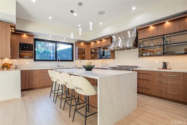 kitchen featuring pendant lighting, a kitchen island with sink, wall chimney exhaust hood, light hardwood / wood-style floors, and a breakfast bar area