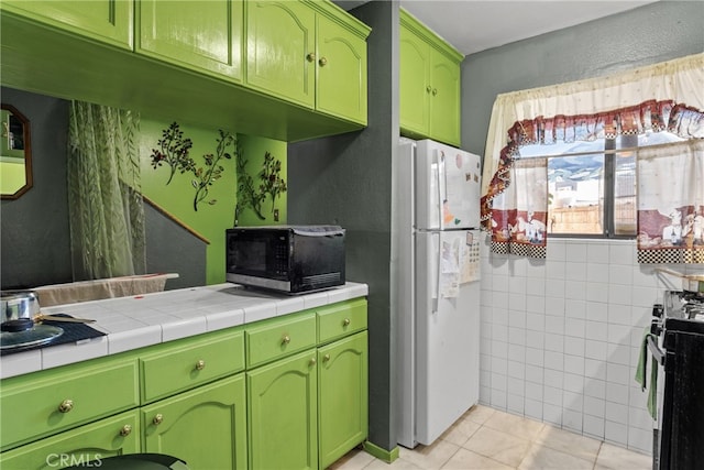 kitchen with white refrigerator, tile countertops, light tile patterned flooring, green cabinetry, and range