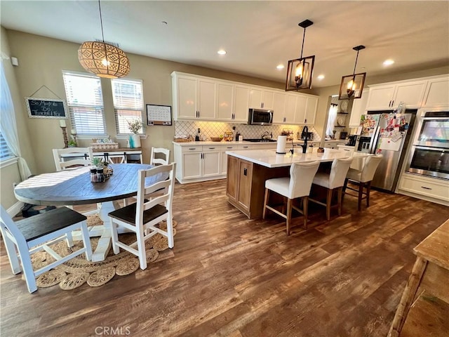 kitchen with pendant lighting, appliances with stainless steel finishes, white cabinetry, an island with sink, and dark hardwood / wood-style floors