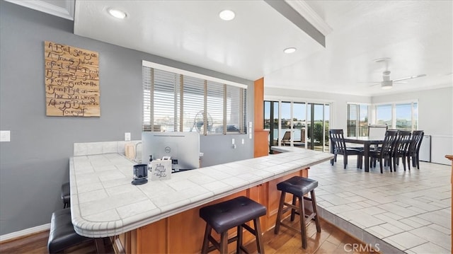 kitchen with ceiling fan, light hardwood / wood-style flooring, ornamental molding, a kitchen bar, and tile countertops