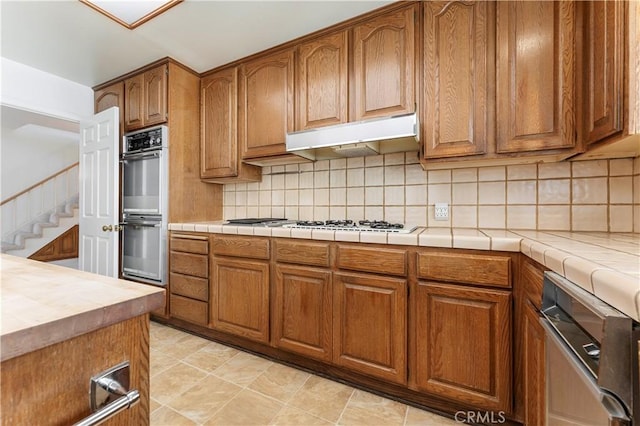 kitchen with under cabinet range hood, multiple ovens, brown cabinetry, and tasteful backsplash