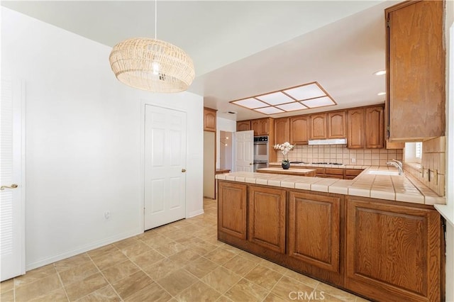 kitchen featuring brown cabinets, double oven, under cabinet range hood, a peninsula, and tasteful backsplash