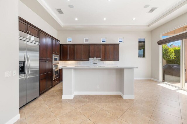 kitchen featuring light tile patterned floors, built in appliances, tasteful backsplash, a tray ceiling, and a center island with sink