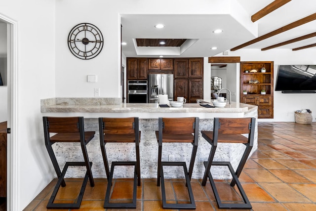 kitchen with beam ceiling, kitchen peninsula, appliances with stainless steel finishes, a tray ceiling, and a breakfast bar