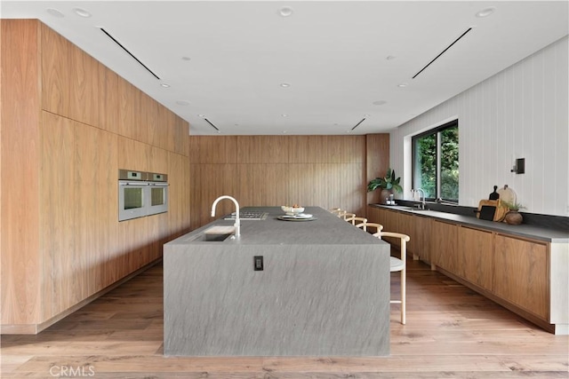 kitchen with wood walls, stainless steel oven, a spacious island, sink, and light hardwood / wood-style flooring