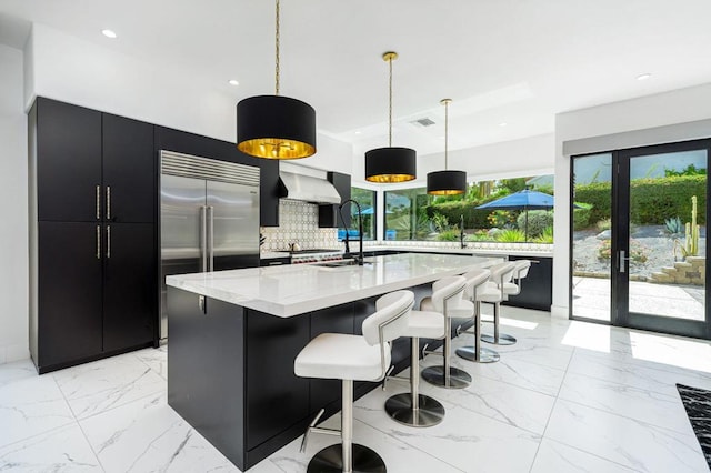 kitchen featuring decorative backsplash, an island with sink, a kitchen bar, decorative light fixtures, and stainless steel built in refrigerator