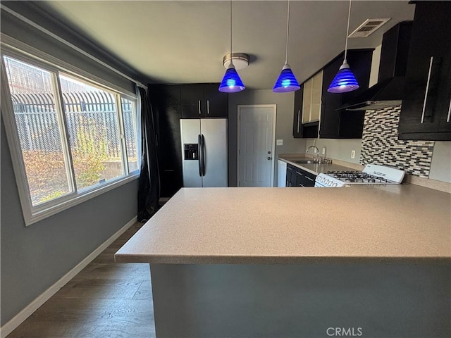 kitchen with range, stainless steel refrigerator with ice dispenser, decorative backsplash, hanging light fixtures, and wall chimney exhaust hood