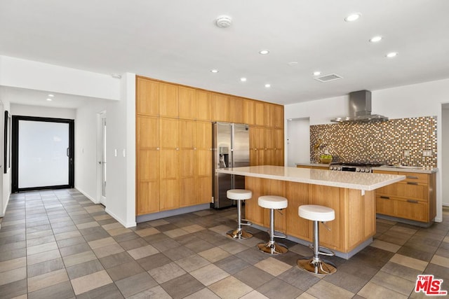 kitchen featuring stainless steel refrigerator with ice dispenser, backsplash, a kitchen island, wall chimney exhaust hood, and a breakfast bar