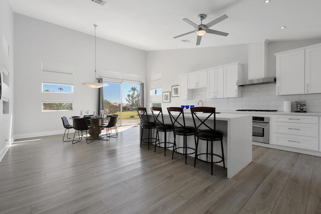 kitchen featuring a kitchen island with sink, wall chimney range hood, pendant lighting, wood-type flooring, and white cabinets