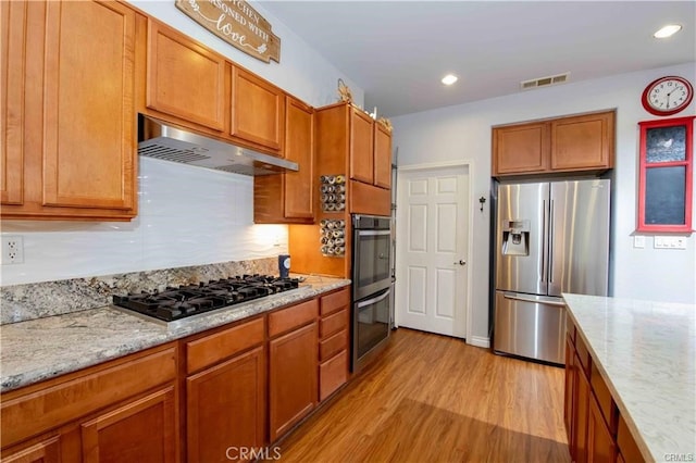 kitchen with appliances with stainless steel finishes, light wood-type flooring, light stone counters, and extractor fan