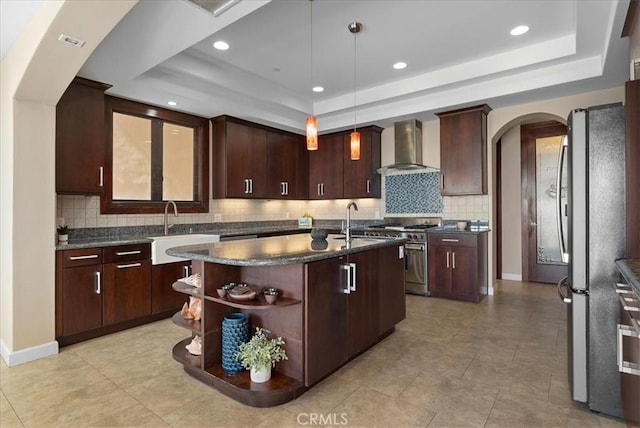 kitchen featuring a raised ceiling, wall chimney exhaust hood, pendant lighting, and a center island with sink