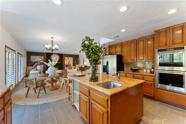 kitchen with sink, a kitchen island with sink, stainless steel appliances, light stone countertops, and decorative light fixtures