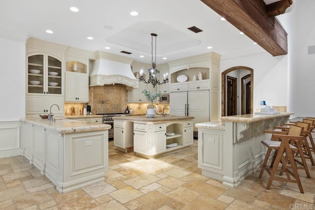 kitchen featuring a kitchen island with sink, hanging light fixtures, high quality appliances, light stone counters, and custom range hood