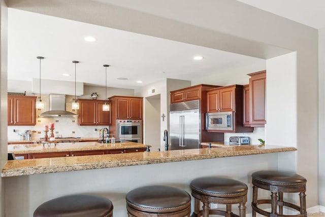 kitchen featuring built in appliances, wall chimney exhaust hood, decorative light fixtures, light stone counters, and kitchen peninsula