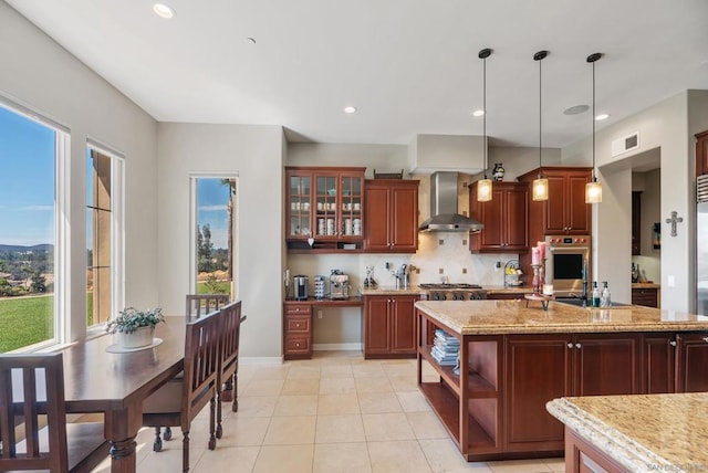 kitchen with wall chimney range hood, decorative backsplash, light stone countertops, light tile patterned floors, and decorative light fixtures