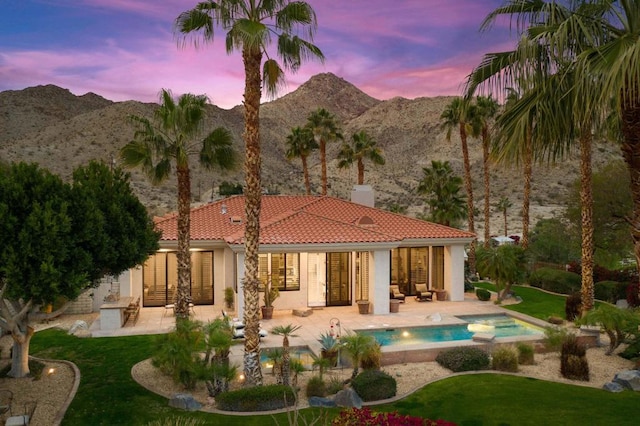 back house at dusk featuring a mountain view, a swimming pool with hot tub, a patio, and a lawn