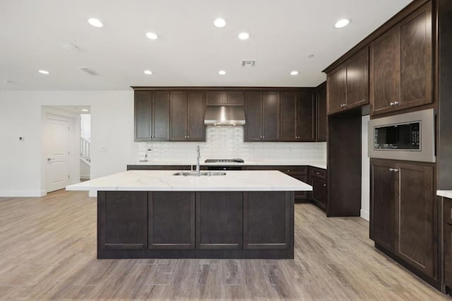 kitchen with light wood-type flooring, light stone counters, sink, stainless steel microwave, and a center island with sink