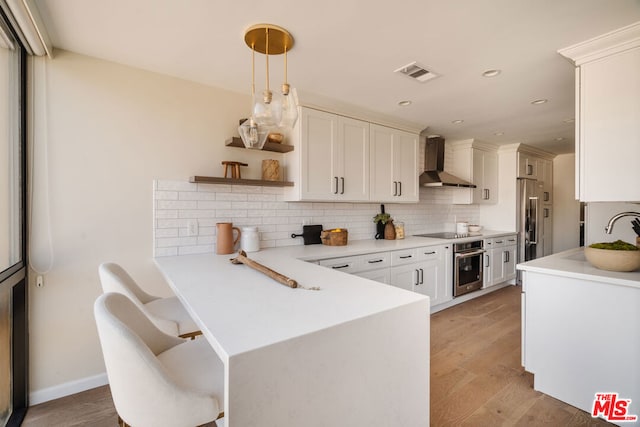 kitchen featuring wall chimney exhaust hood, kitchen peninsula, light hardwood / wood-style flooring, appliances with stainless steel finishes, and backsplash