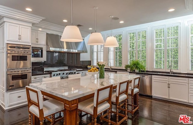 kitchen featuring wall chimney range hood, appliances with stainless steel finishes, hanging light fixtures, dark stone countertops, and crown molding